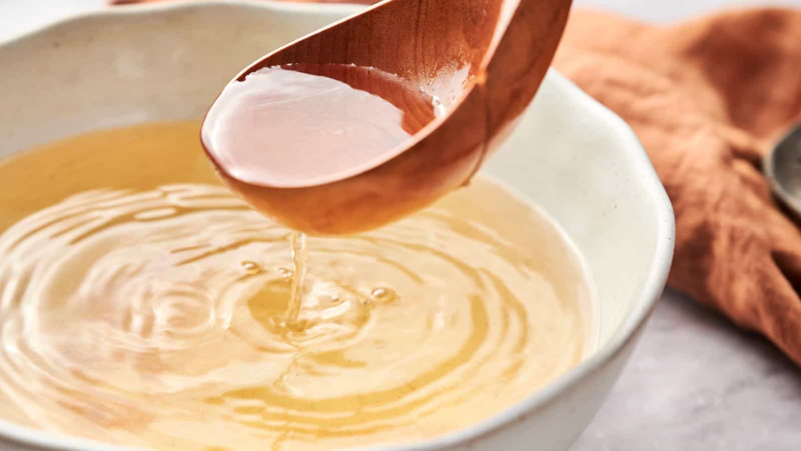 A wooden ladle pouring fish sauce into a white ceramic bowl, with ripples visible on the liquid surface. A brown napkin is partially visible in the background.