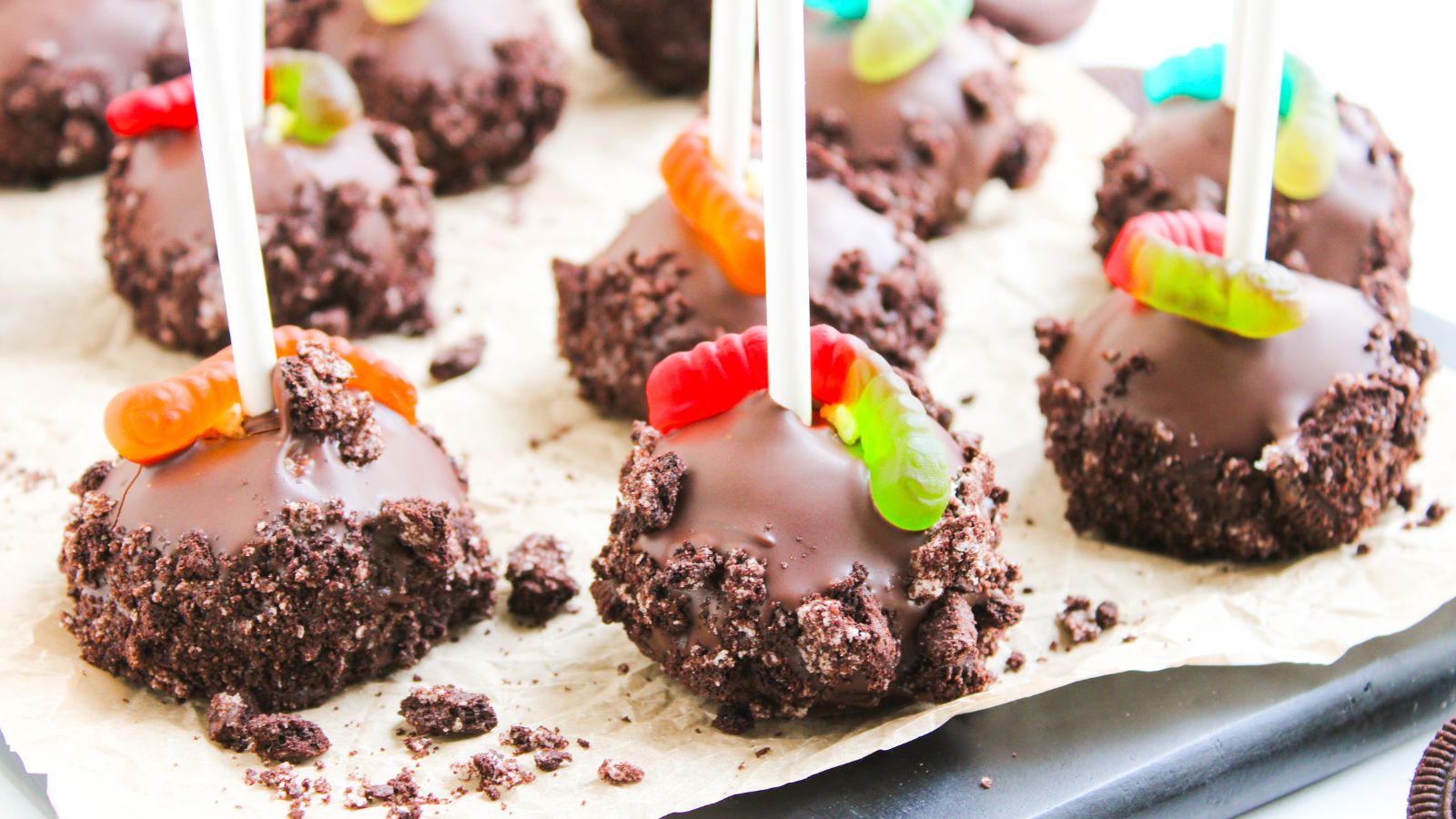 Chocolate cake pops covered in crushed cookies, each with a gummy worm on top, displayed on parchment paper.