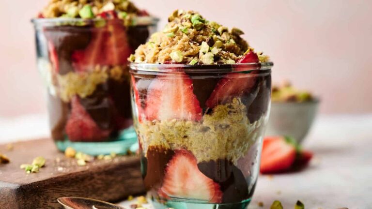 Two glass cups filled with layers of sliced strawberries, chocolate pudding, oats, and chopped pistachios, set on a table with a spoon and scattered nuts.