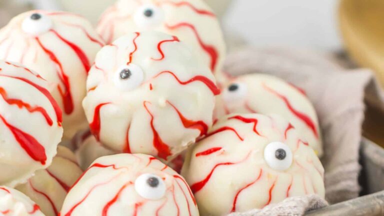 Round white treats decorated with red icing to look like bloodshot eyeballs, featuring a single candy eye on each, arranged in a pile.