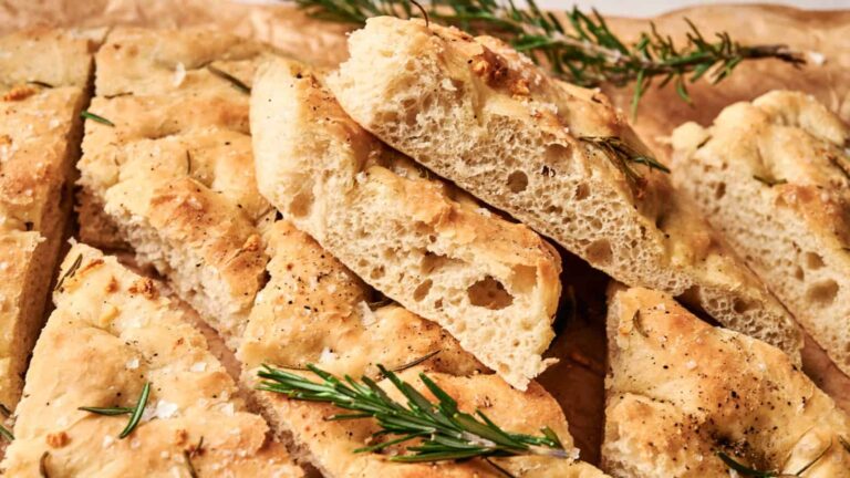 Slices of golden brown focaccia bread topped with rosemary and coarse salt, arranged on parchment paper.