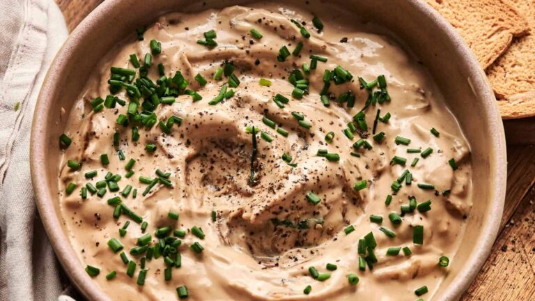 A bowl of creamy dip topped with chopped chives and ground black pepper, with toast slices visible on the side.