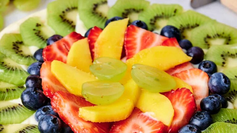 A fruit platter with sliced kiwi, strawberries, mango, blueberries, and green grapes arranged in a circular pattern.