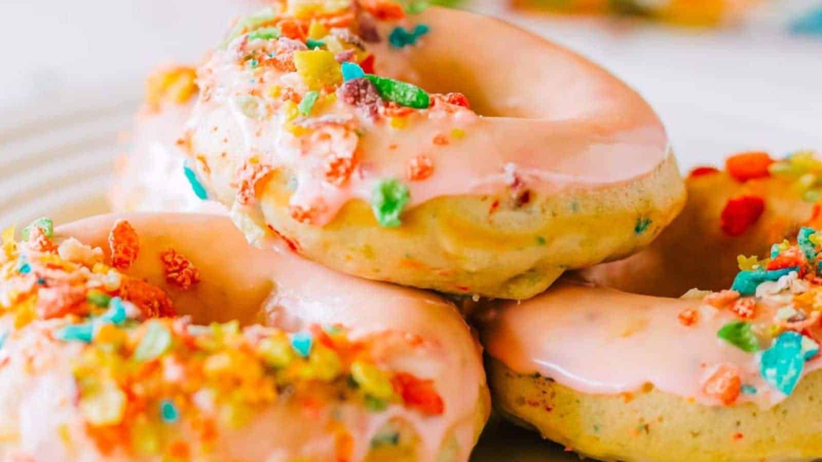 Three donuts with pink frosting and colorful Fruity Pebbles cereal pieces on top, stacked on a white plate.
