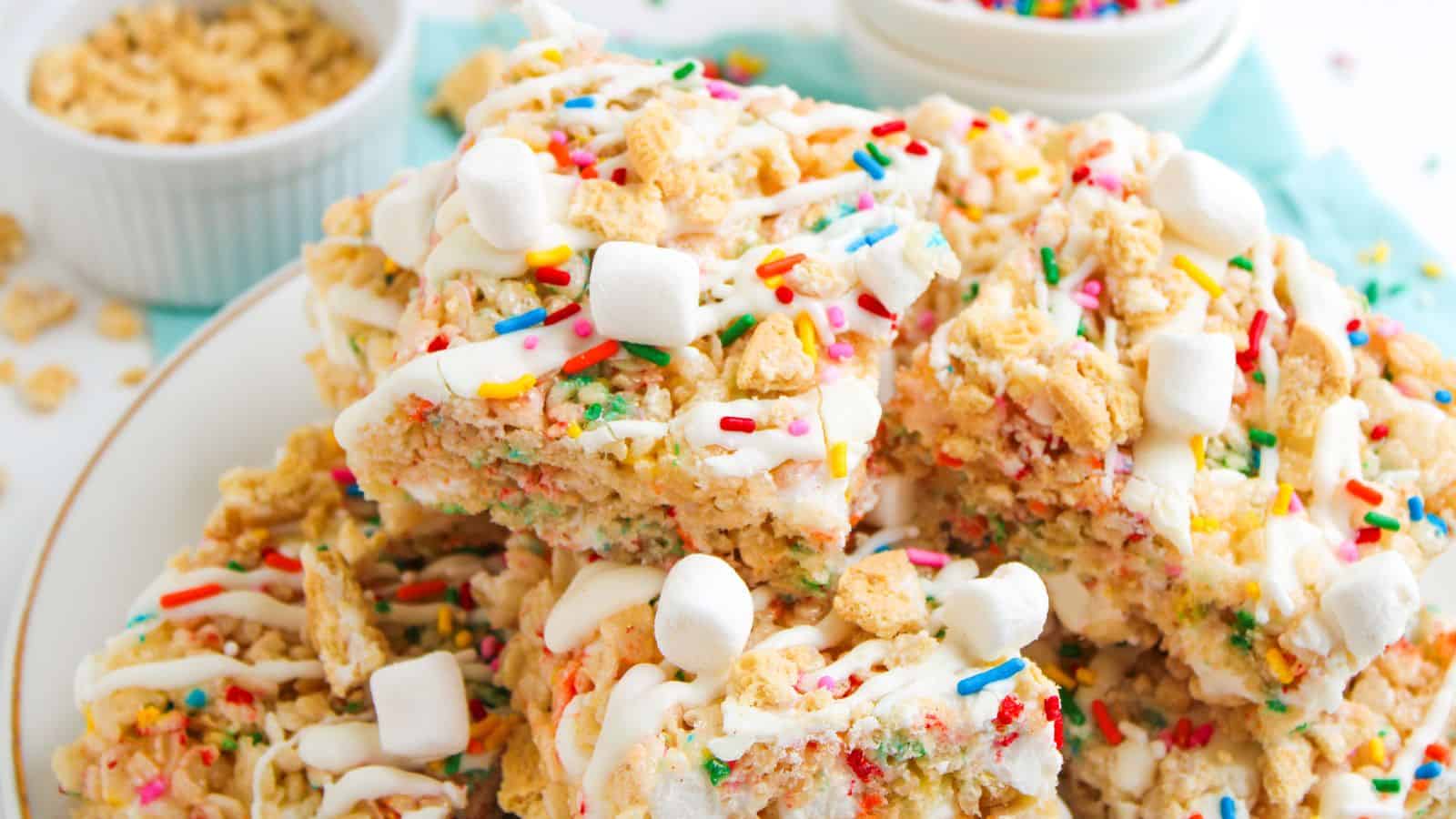 A plate of rice crispy treats topped with white icing, mini marshmallows, and colorful sprinkles, with bowls of cereal and sprinkles in the background.