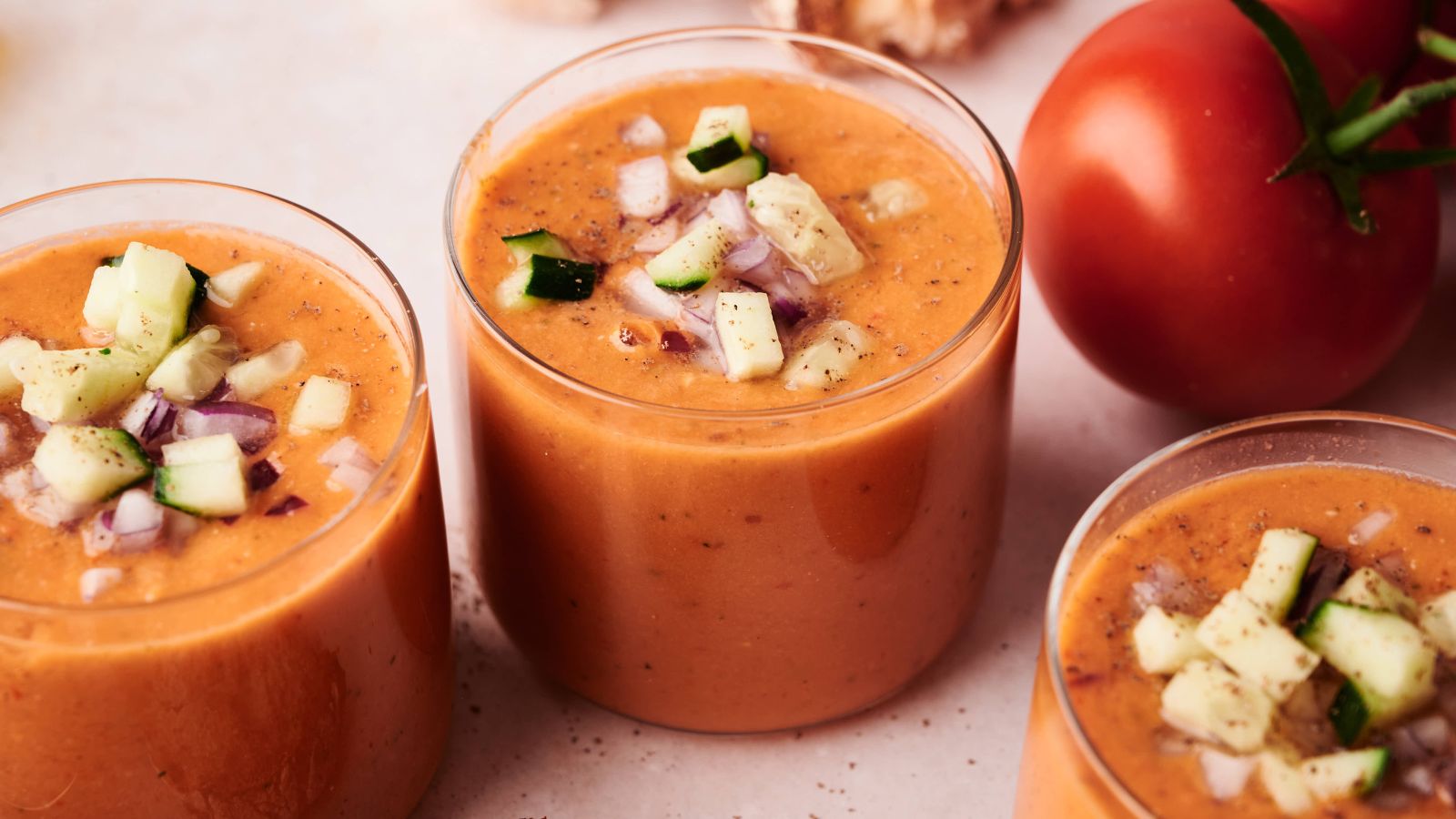 Three glasses of gazpacho soup topped with chopped cucumber and red onion, with a whole tomato in the background on a light surface.