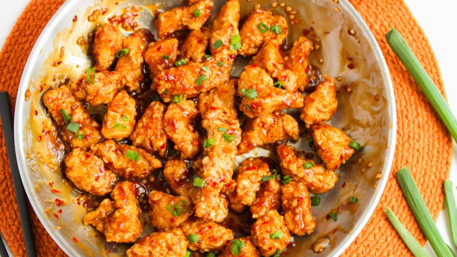Stir-fried chicken pieces in a spicy sauce, garnished with chopped green onions, served in a round metal dish on an orange placemat.