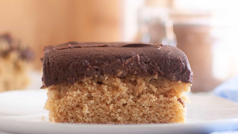A close-up of a single square piece of cake with a thick layer of chocolate frosting on top, served on a white plate.