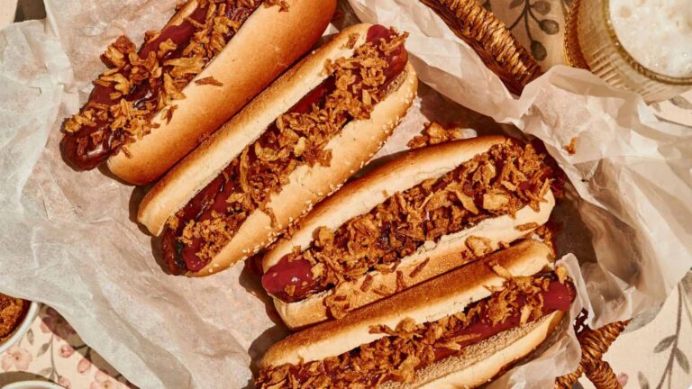Four hot dogs in buns topped with crispy fried onions are arranged on parchment paper in a basket.