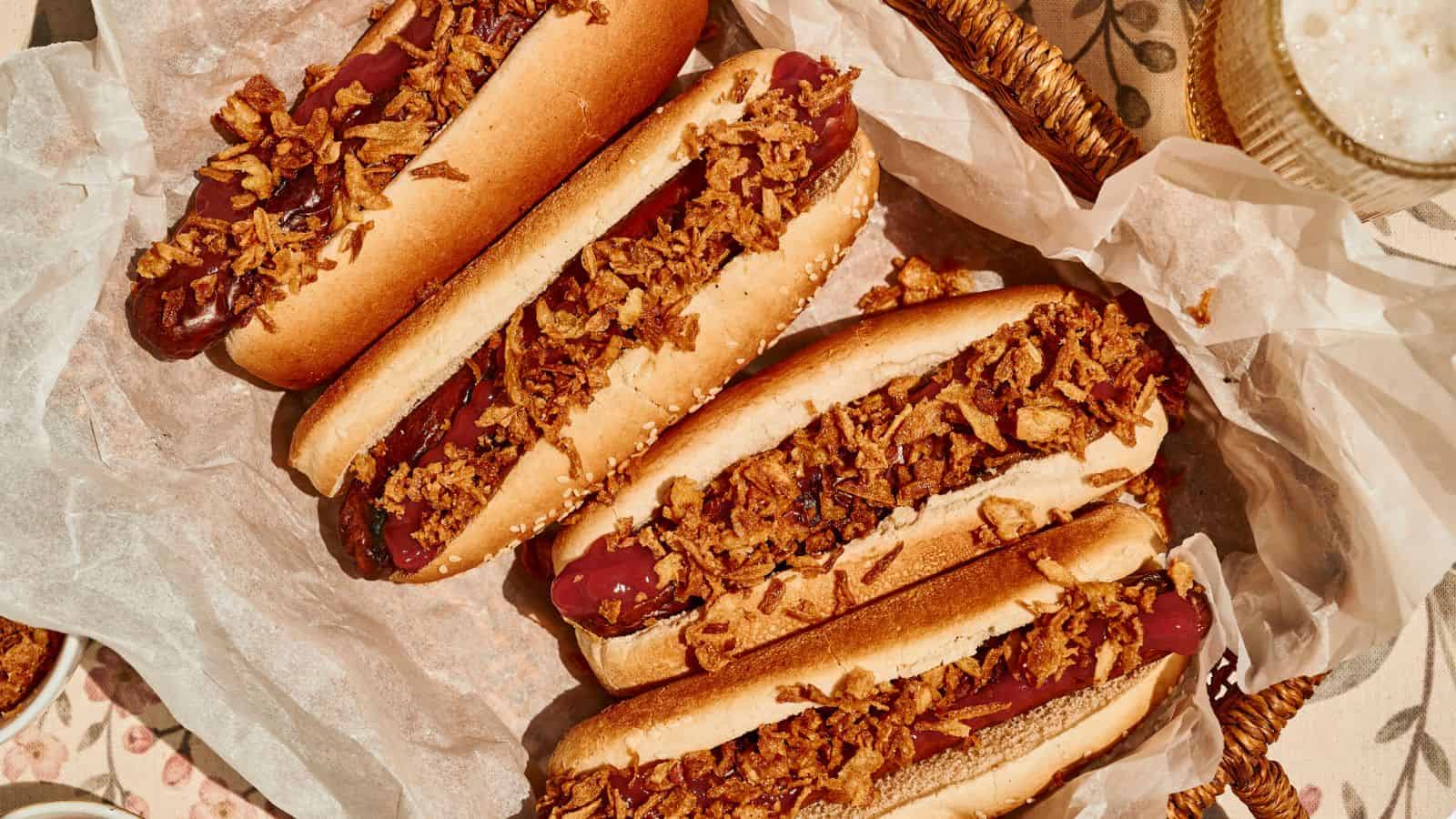 Four hot dogs in buns topped with crispy fried onions are arranged on parchment paper in a basket.