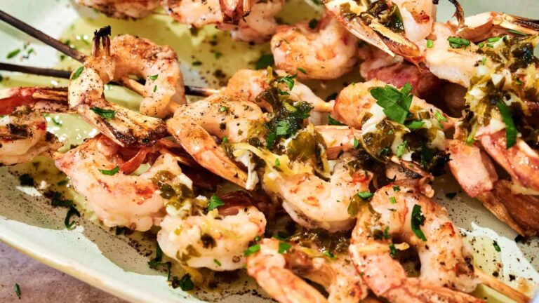 Grilled shrimp skewers garnished with chopped herbs and drizzled with a light sauce, served on a white plate.