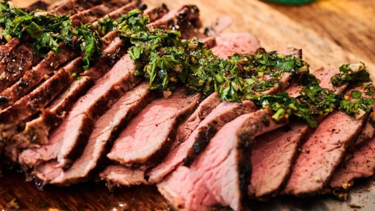 Sliced grilled steak arranged on a wooden board, topped with chopped fresh herbs and green sauce.