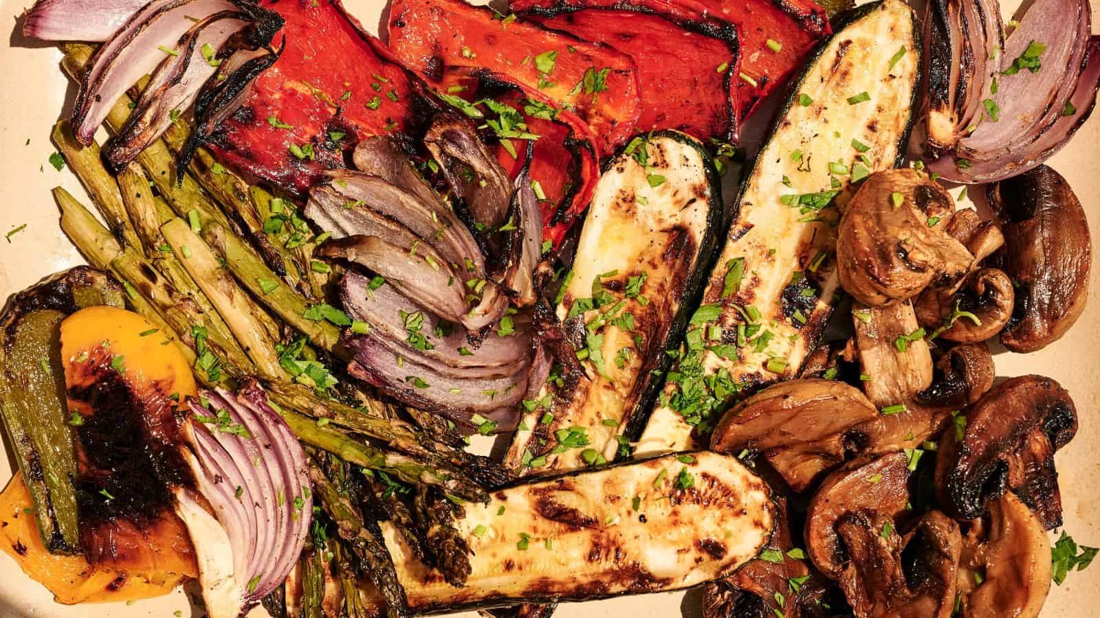 A platter of grilled vegetables including zucchini, mushrooms, asparagus, bell peppers, and red onions, garnished with chopped herbs.
