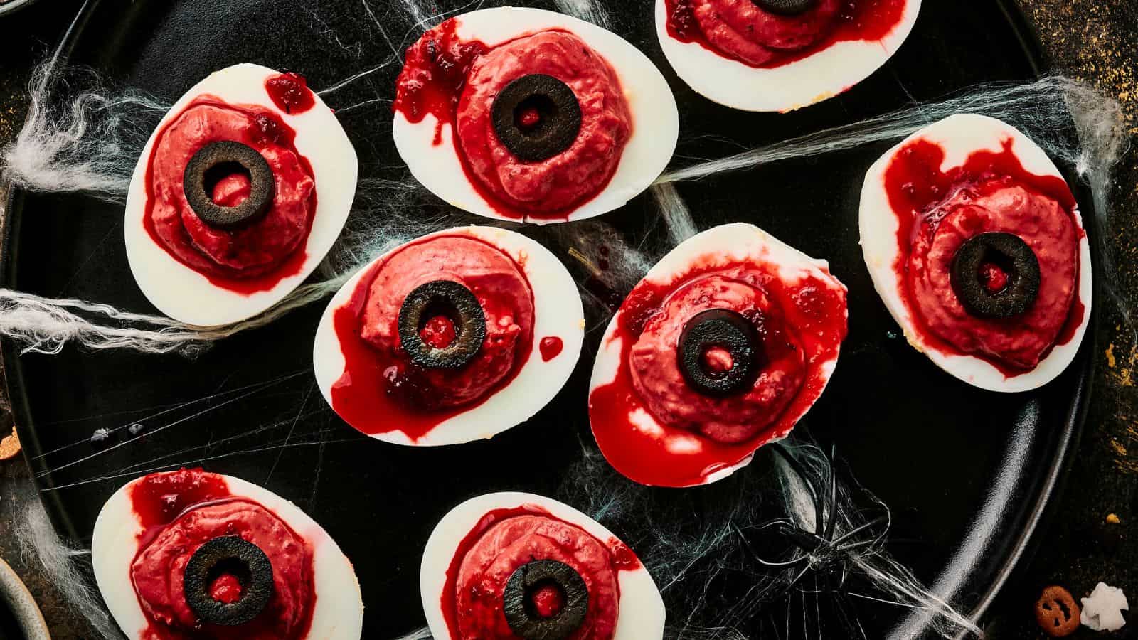 A plate of Halloween deviled eggs, halved and adorned with red sauce and black olives to resemble eerie eyeballs, sits on a dark background with cobwebs.
