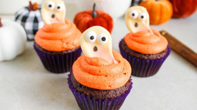 Three chocolate cupcakes with orange frosting are topped with white ghost decorations featuring candy eyes and open mouths. Several decorative pumpkins are in the background.