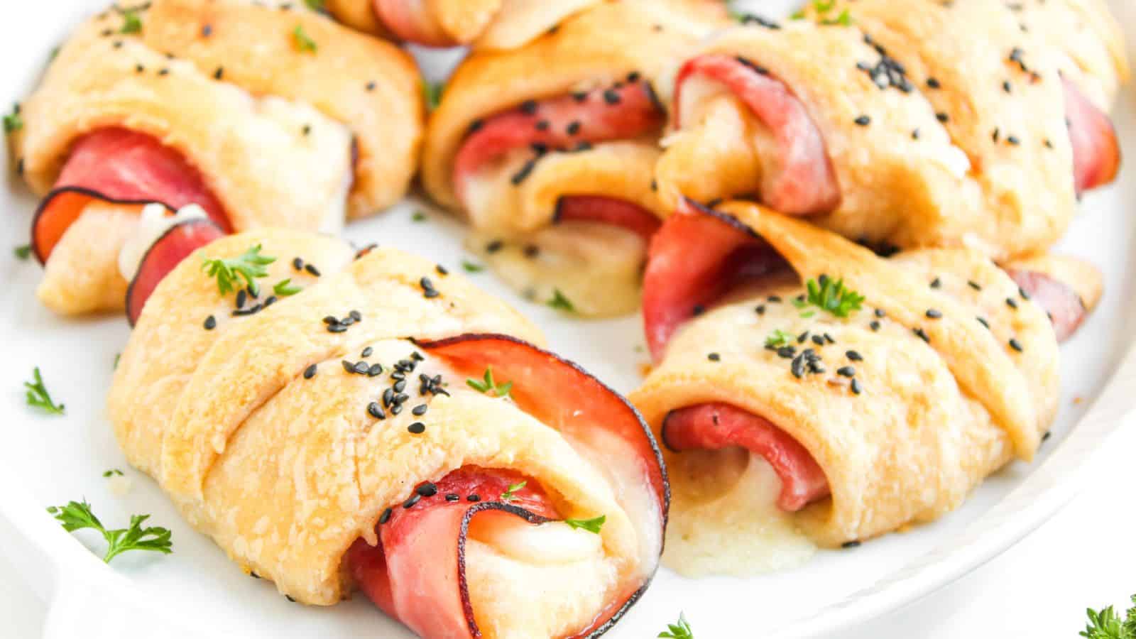 A plate of baked crescent rolls filled with slices of ham and melted cheese, topped with black sesame seeds and garnished with parsley.
