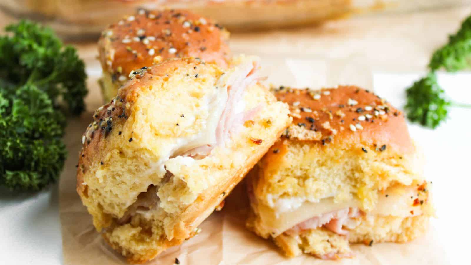 Two ham and cheese sliders on seeded buns are placed on parchment paper. Bright green parsley is visible in the background.