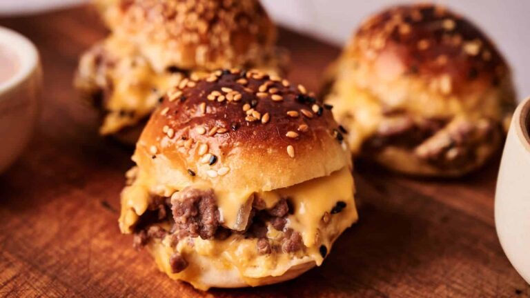 Three sesame seed buns filled with melted cheese and ground beef are arranged on a wooden board.