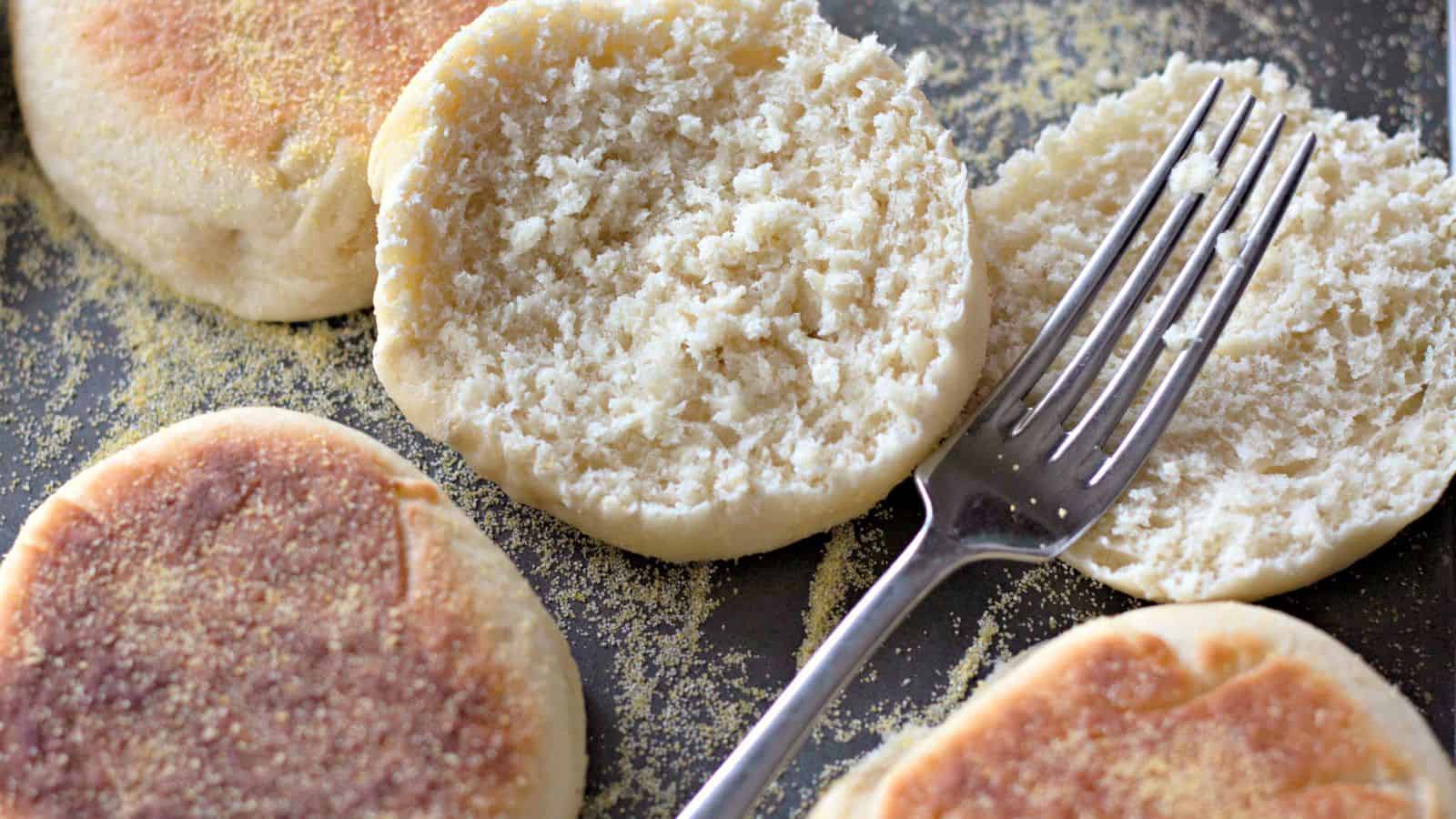 Several English muffins, one split open with a fork, rest on a baking sheet dusted with cornmeal.