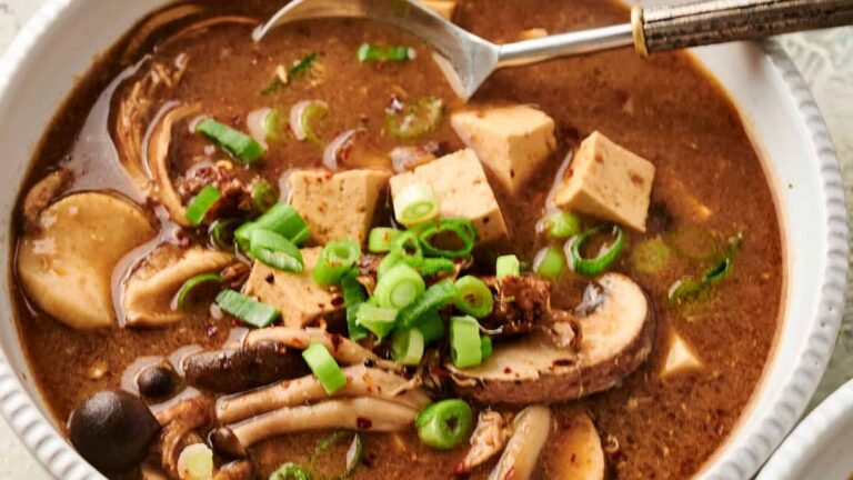 Bowl of hot and sour soup with tofu, mushrooms, sliced green onions, and a spoon.