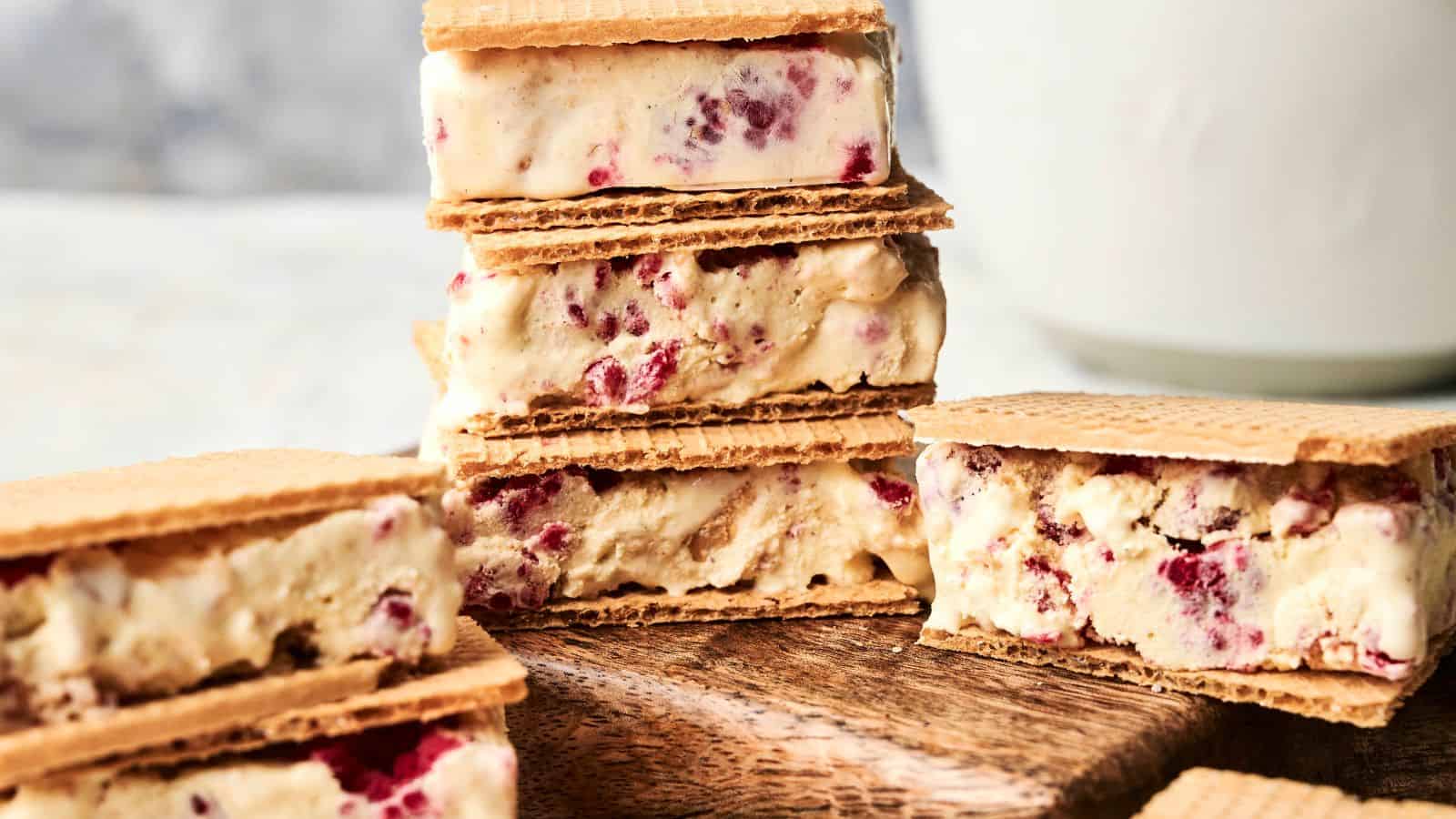 Ice cream sandwiches with wafer cookies and ice cream containing visible berry pieces, arranged on a wooden board with a white background.