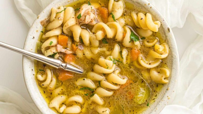 A bowl of rotini pasta soup with pieces of chicken, carrots, celery, and herbs, with a metal spoon resting inside.