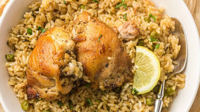 A plate of seasoned rice with two pieces of baked chicken, garnished with a lemon slice and herbs, served with a spoon.