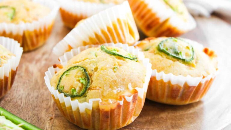Cornbread muffins topped with sliced jalapeños in white paper liners, arranged on a wooden surface.