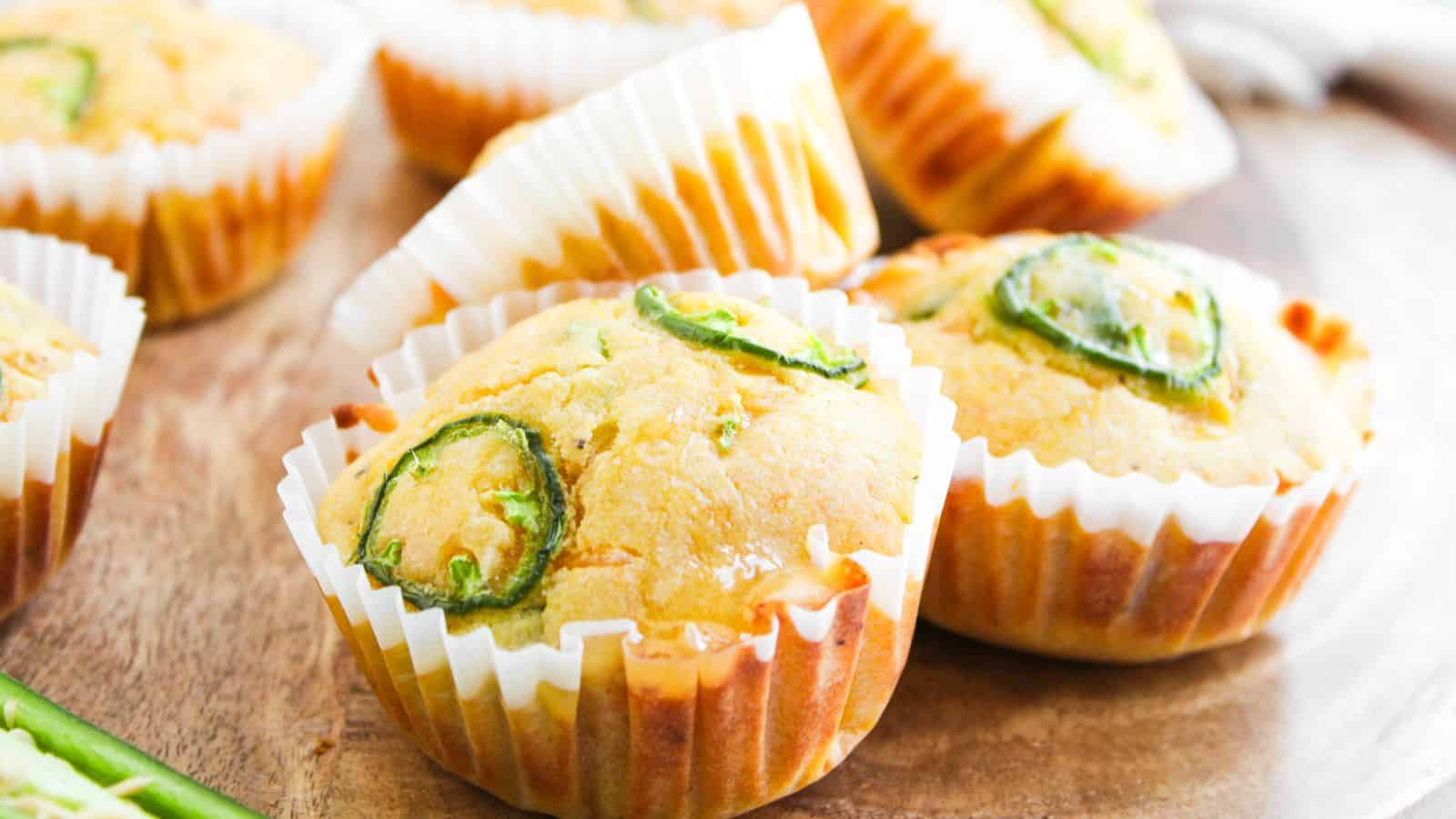 Cornbread muffins topped with sliced jalapeños in white paper liners, arranged on a wooden surface.