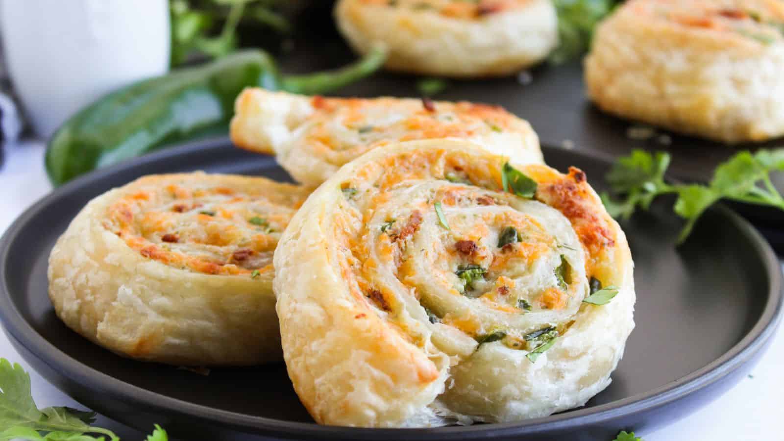Close-up of three spiral pastry rolls on a black plate, filled with herbs and cheese. Green garnish and a jalapeño pepper are in the background.