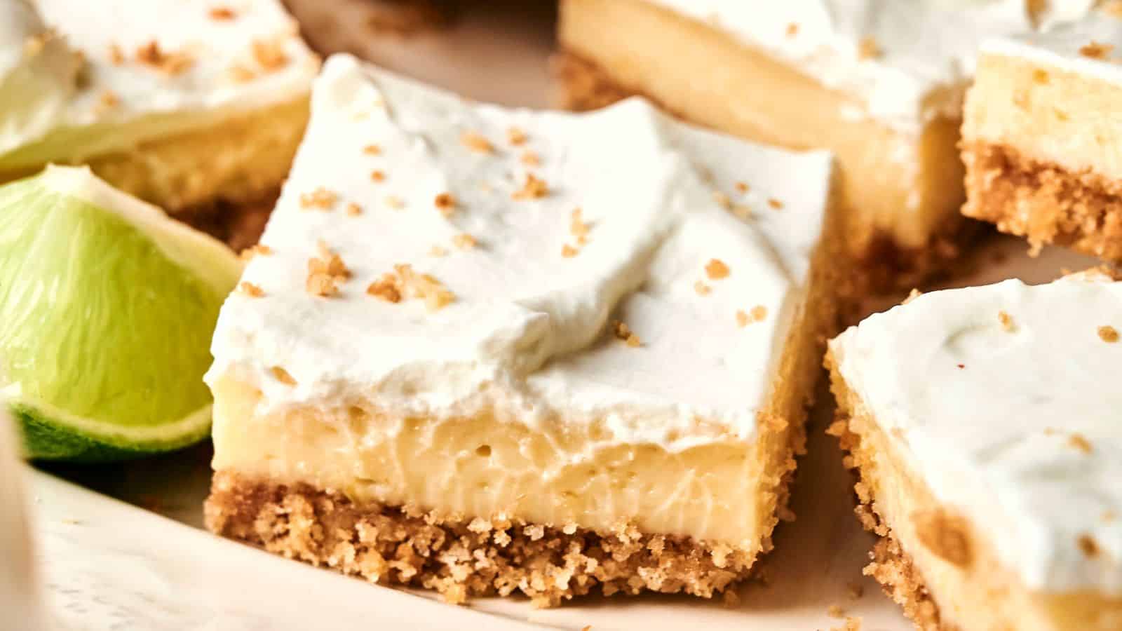 A close-up of dessert bars with a crumbly crust, creamy filling, and whipped topping, garnished with lime slices in the background.