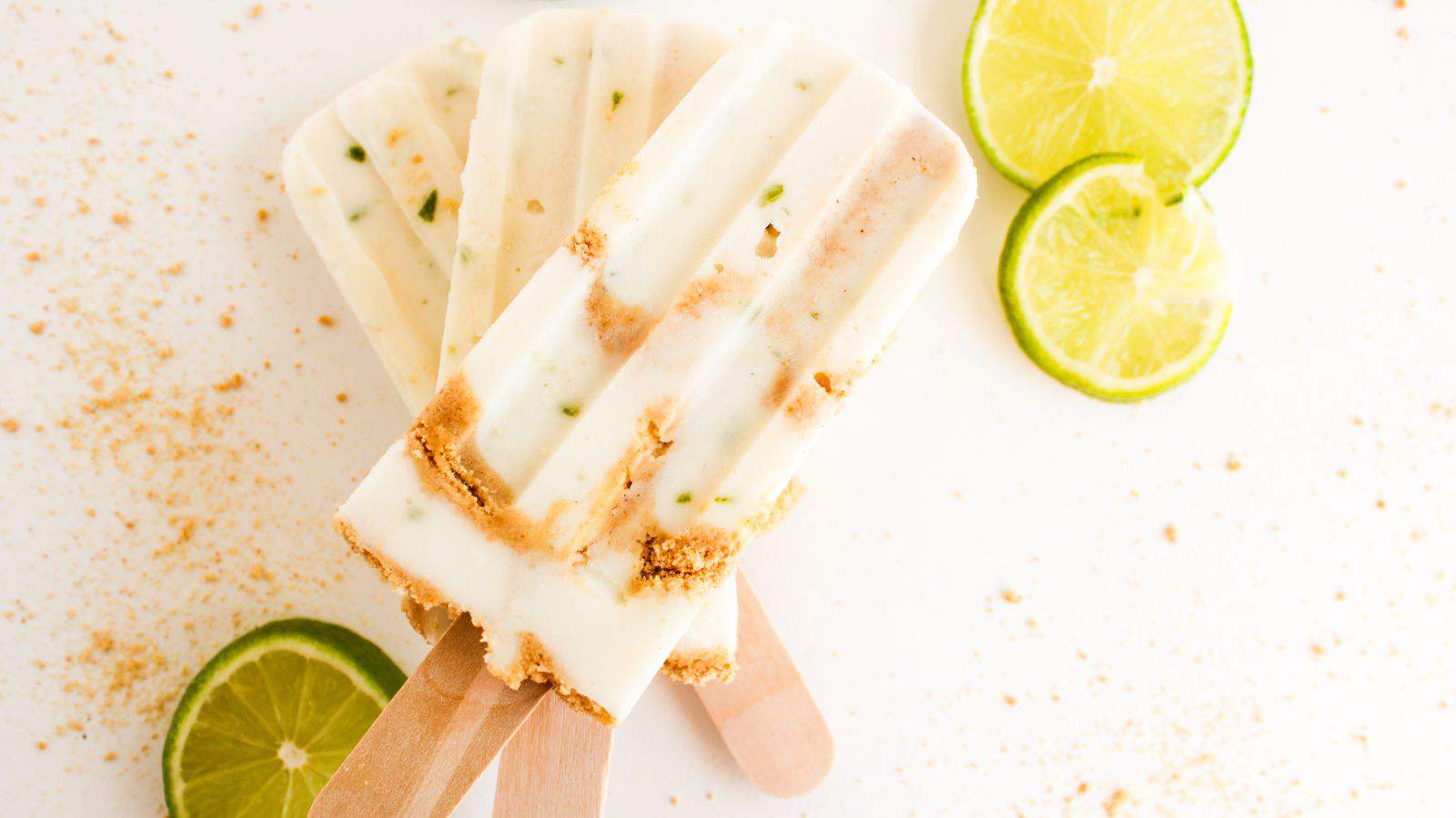 Three creamy popsicles with lime slices and crumbs scattered around on a white surface.