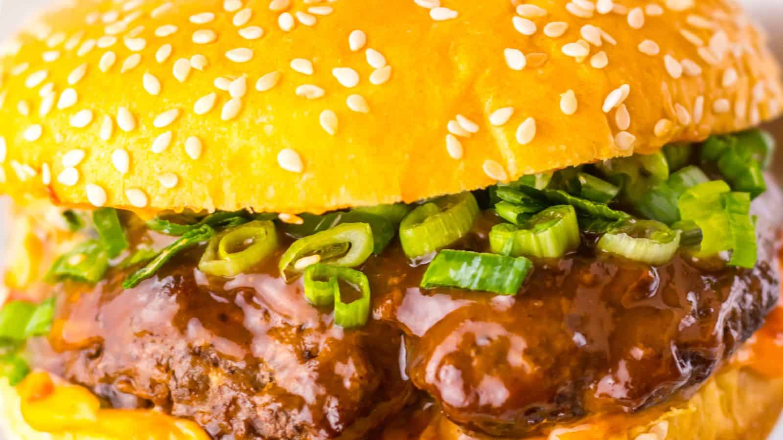 Close-up of a hamburger with a sesame seed bun, beef patty, green onions, and rich brown sauce.
