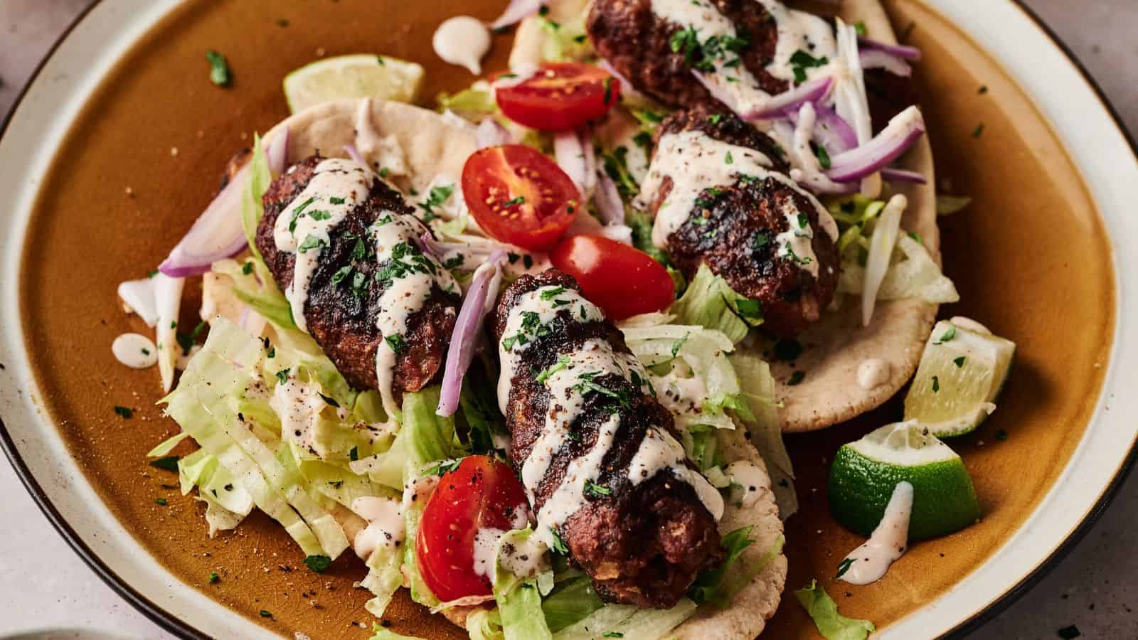 Three grilled kebabs on flatbread, topped with shredded lettuce, cherry tomatoes, red onion, chopped herbs, and a drizzle of white sauce, served with lime wedges.