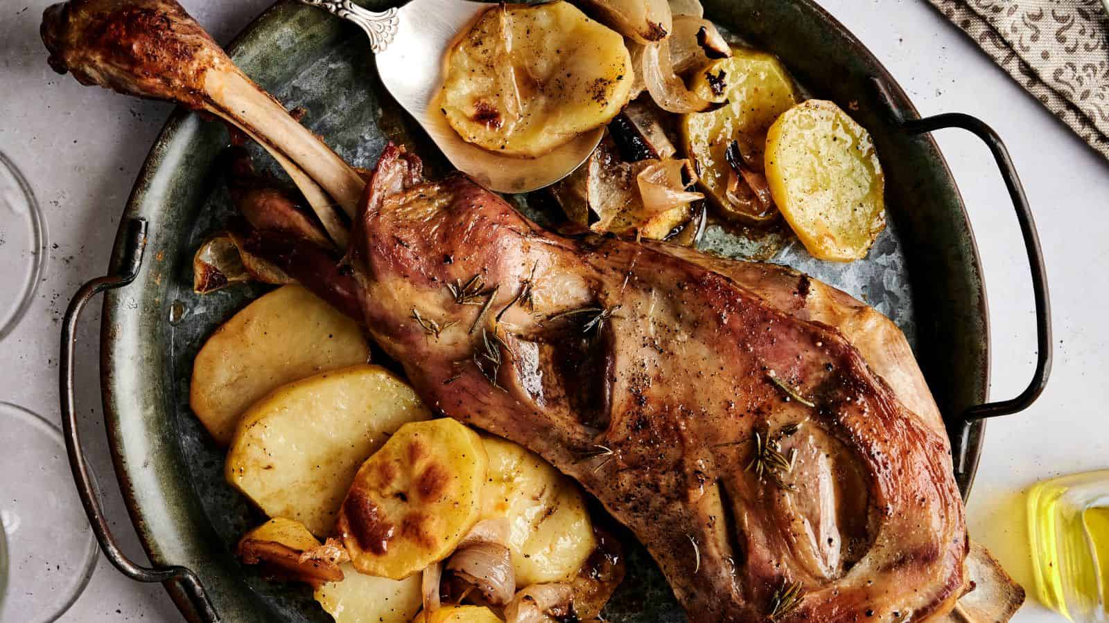 Roasted lamb leg on a platter with sliced potatoes, lemon, and herbs. A knife is placed beside the dish.