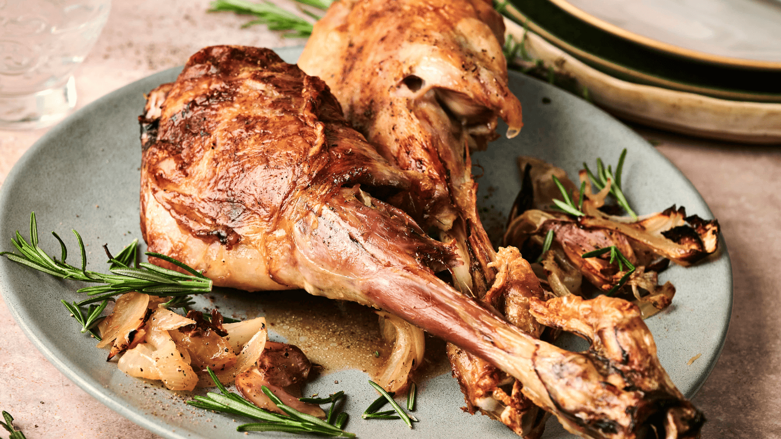 Two roasted lamb shanks on a plate with rosemary and caramelized onions, with stacked plates in the background.