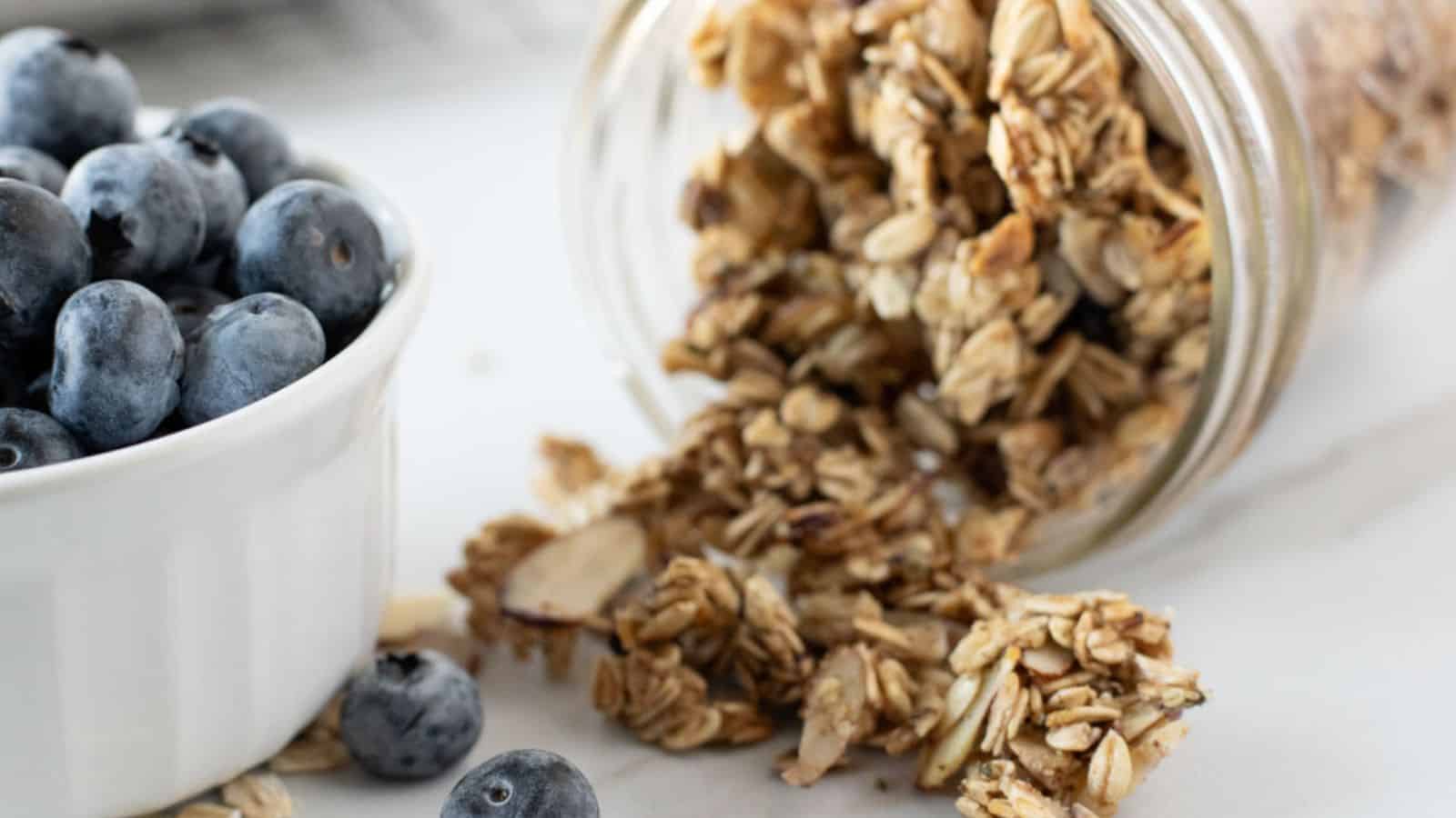 A glass jar of granola spills onto a surface next to a white bowl filled with blueberries.