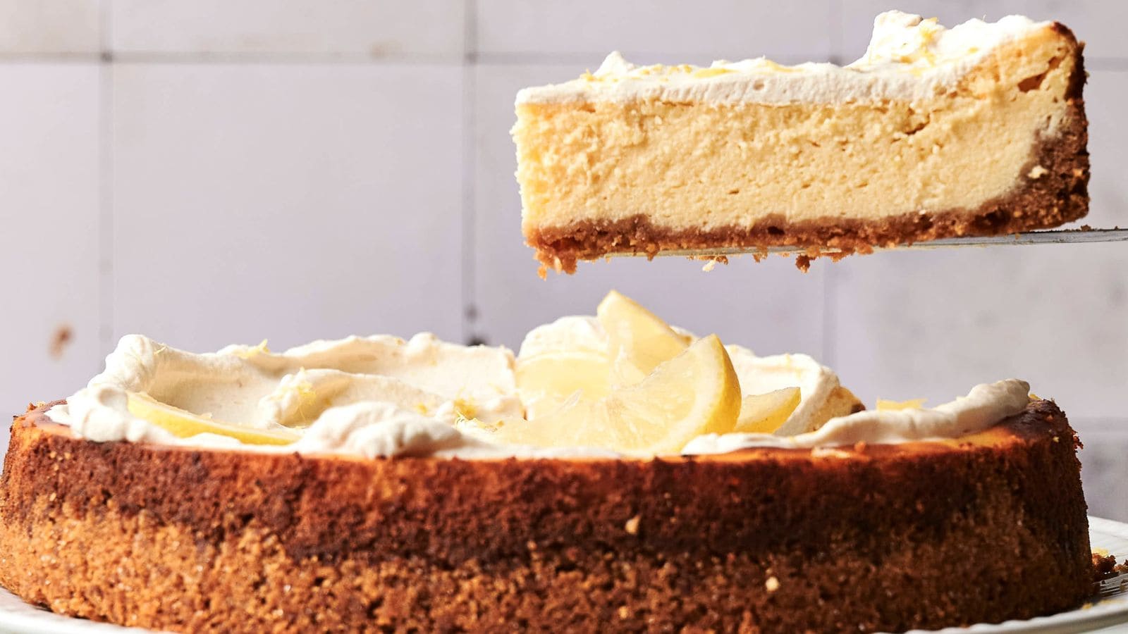 A slice of lemon cheesecake with a graham cracker crust is being lifted from the whole cake, which is topped with whipped cream and lemon slices.