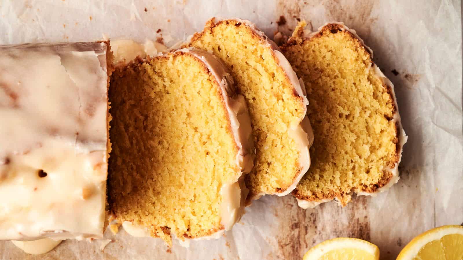 Sliced lemon cake with glaze on parchment paper, next to lemon slices.