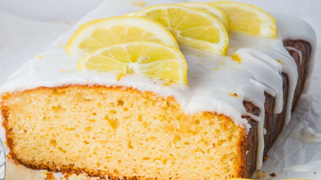 A loaf of lemon pound cake topped with white icing and lemon slices, shown with a sliced end to reveal its texture.