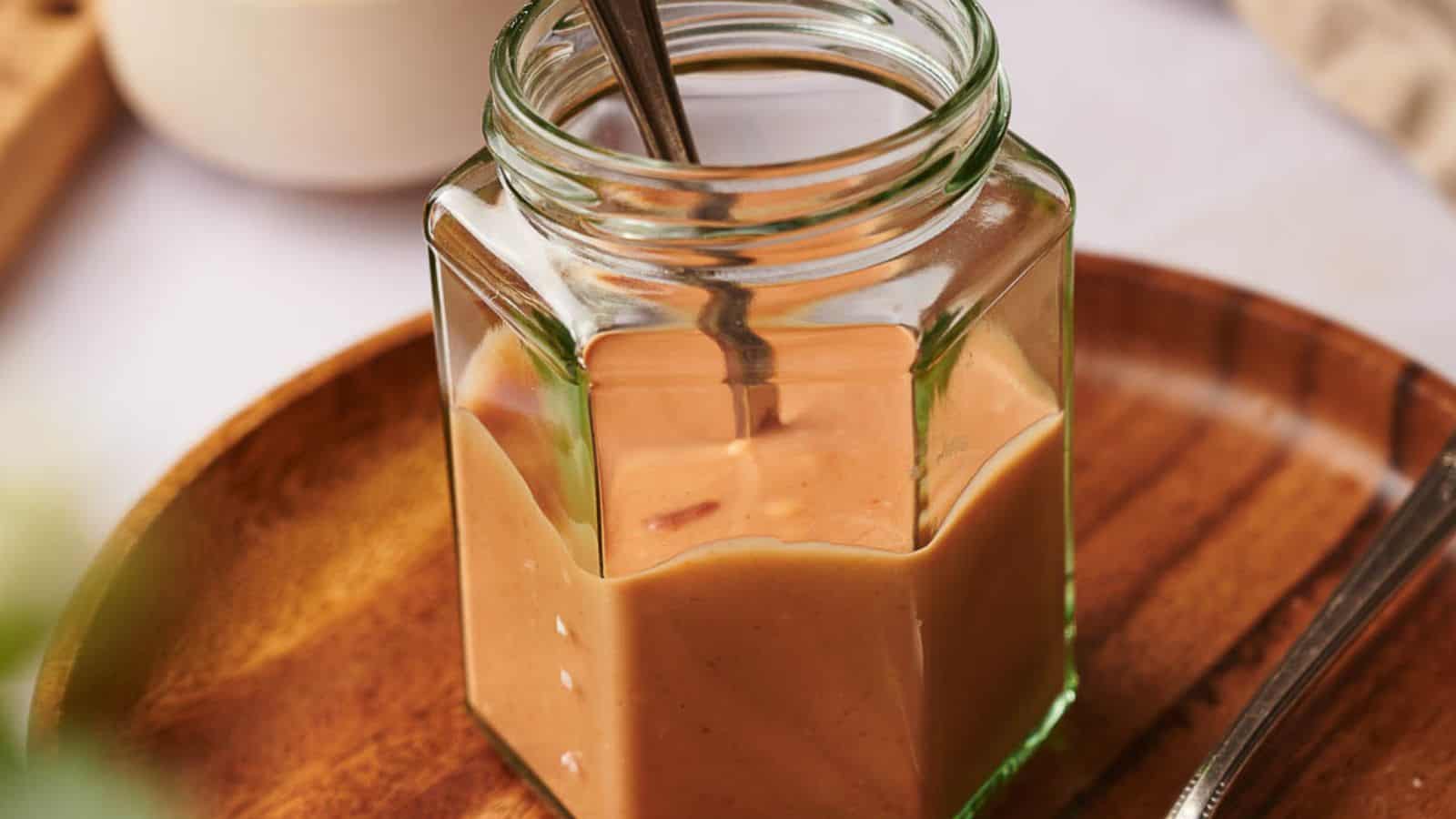 A hexagonal glass jar filled with creamy peanut butter sits on a wooden tray, with a metal spoon inside.