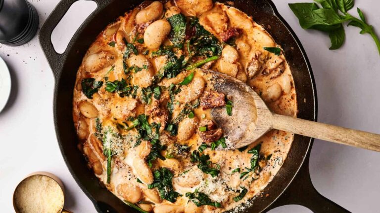 A cast iron skillet filled with creamy beans, spinach, and sun-dried tomatoes, stirred with a wooden spoon, garnished with grated cheese.
