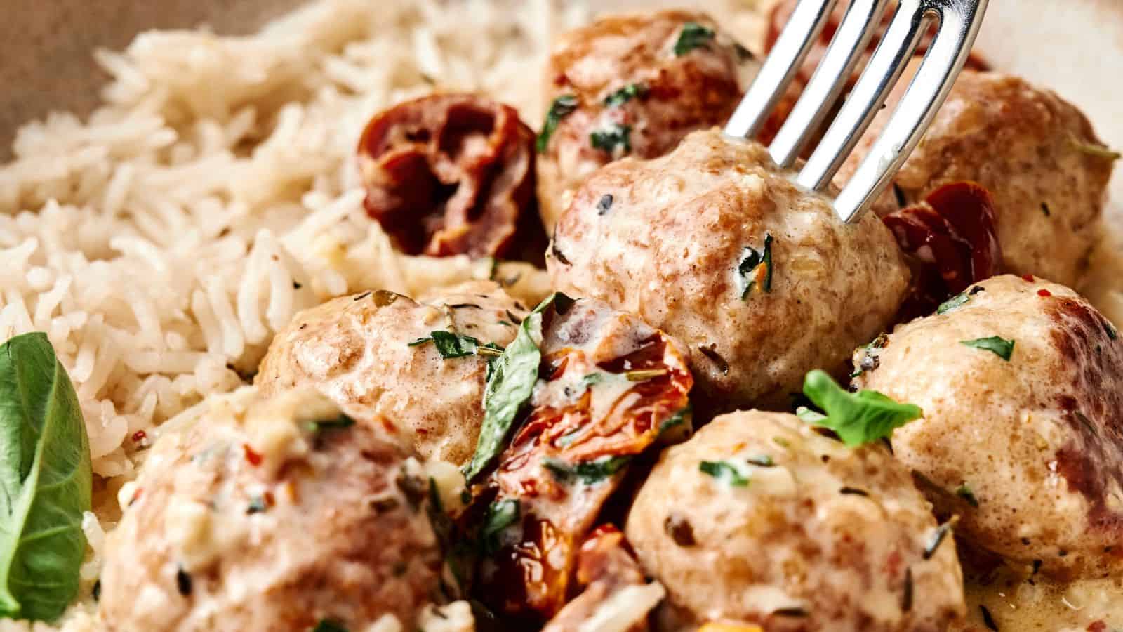 A close-up of meatballs in a creamy sauce with herbs, sun-dried tomatoes, and a side of white rice, with a fork picking up one meatball.