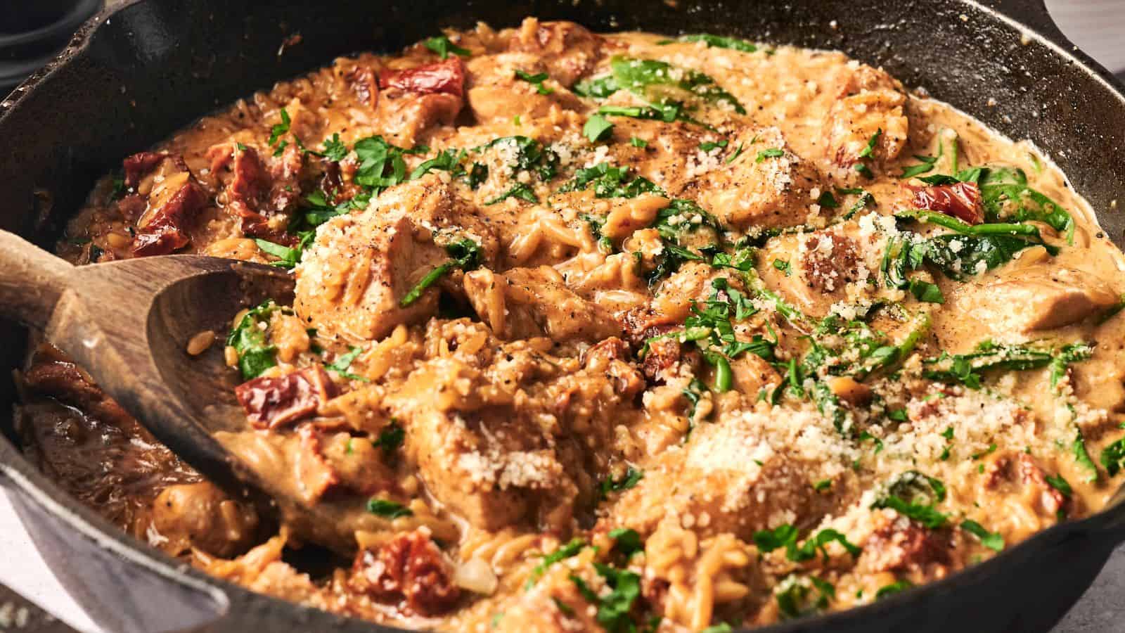 A skillet with creamy chicken pasta featuring spinach, sun-dried tomatoes, and grated cheese, stirred with a wooden spoon.