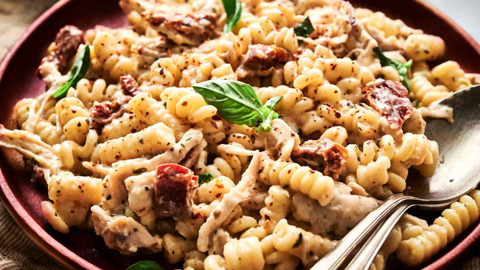 A plate of creamy rotini pasta with shredded chicken, sun-dried tomatoes, herbs, and a spoon placed at the side.