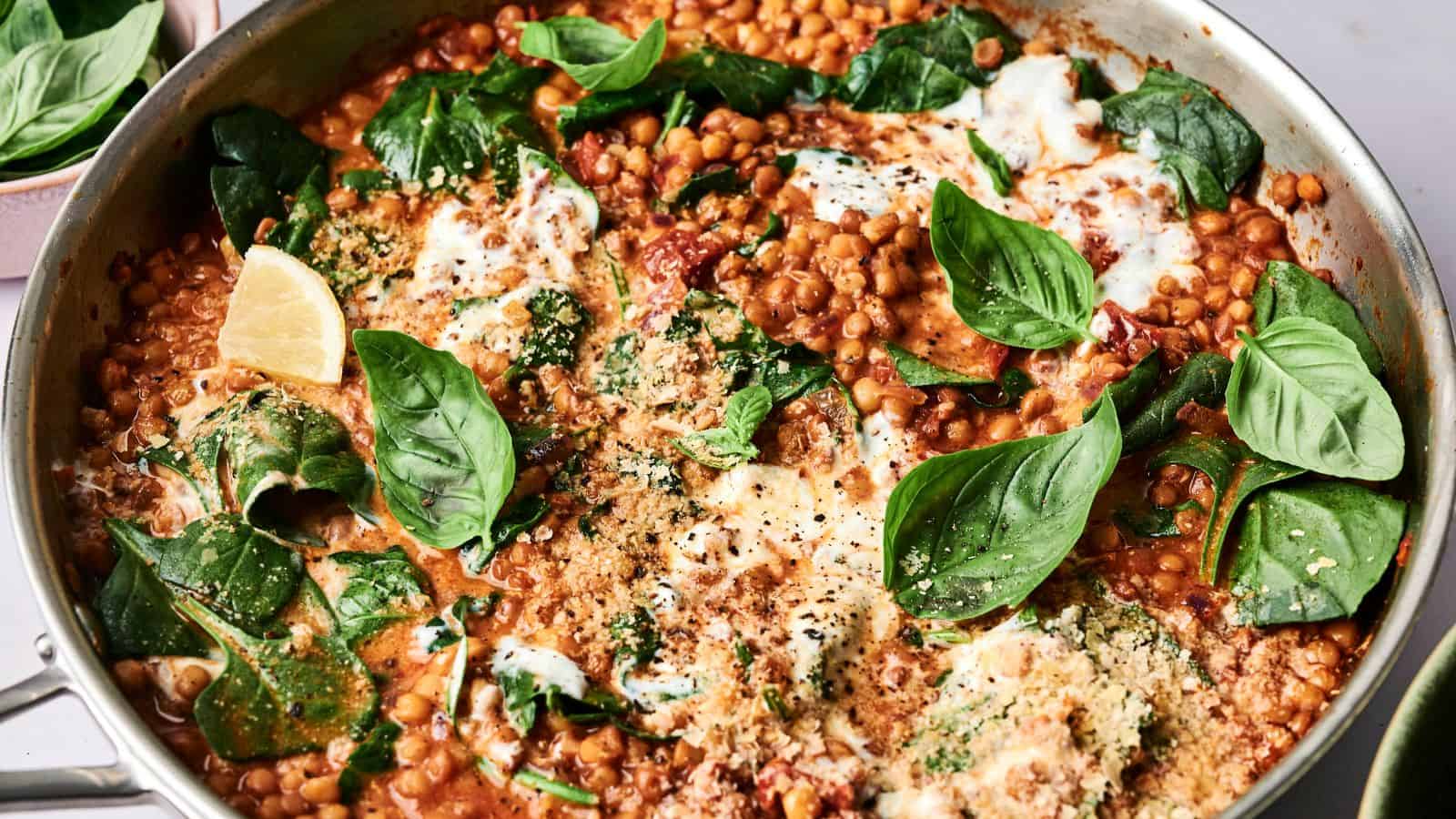 A pan of lentil dish with spinach, fresh basil, tomato sauce, and melted cheese, garnished with lemon wedges.