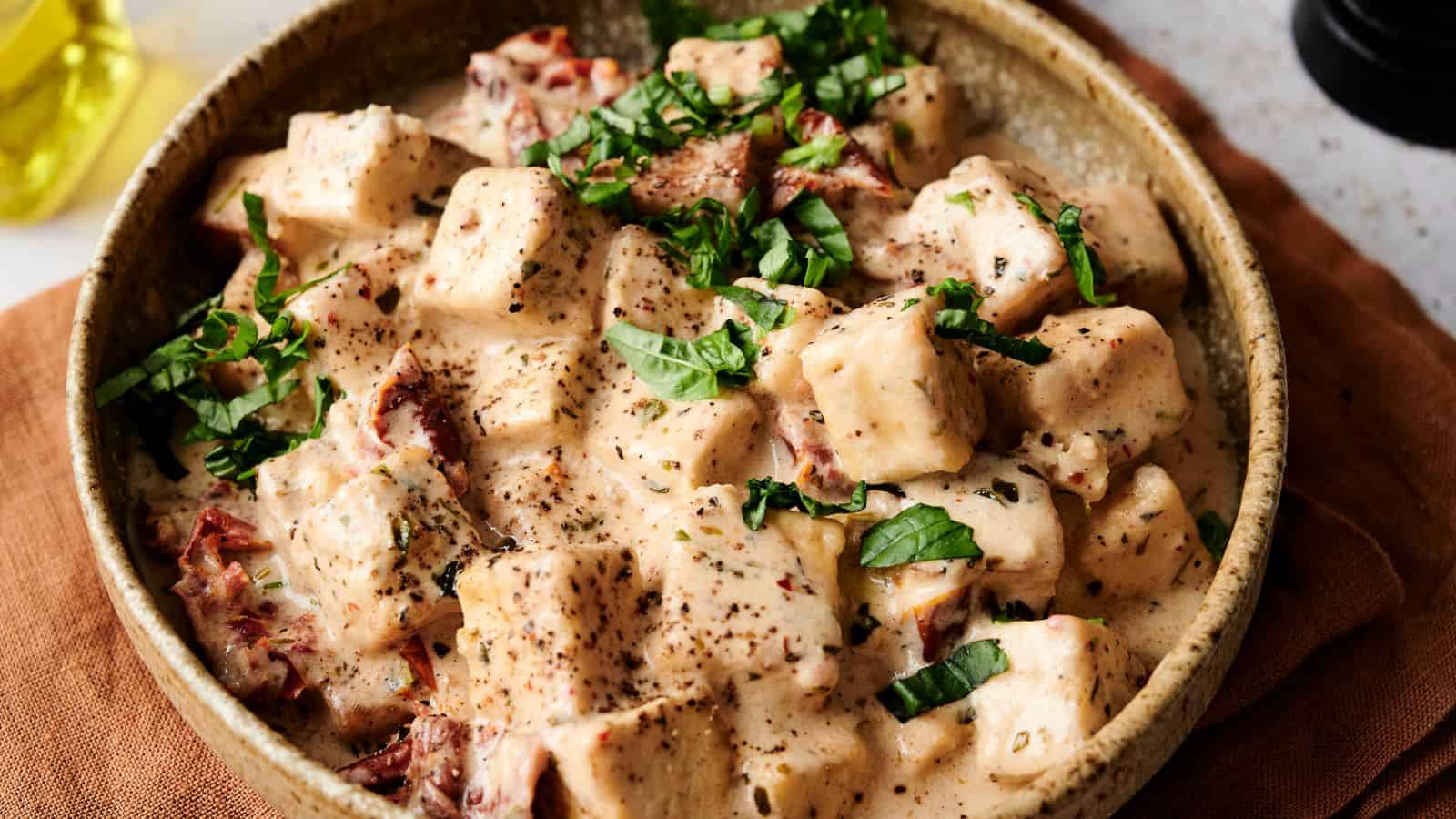 A bowl of creamy pasta salad with cubed noodles and sun-dried tomatoes, garnished with fresh basil leaves and black pepper, served in a rustic dish on a brown cloth.