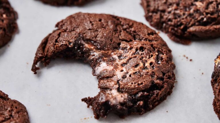 A chocolate cookie with a bite taken out, showing a gooey, melty center, rests on a white surface among other cookies.