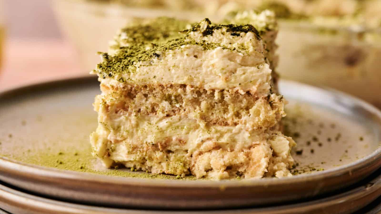 A close-up of a slice of tiramisu topped with green matcha powder, served on a ceramic plate.