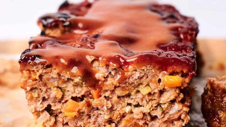 Close-up of a sliced meatloaf topped with a glossy tomato-based sauce, showing visible chunks of vegetables inside.